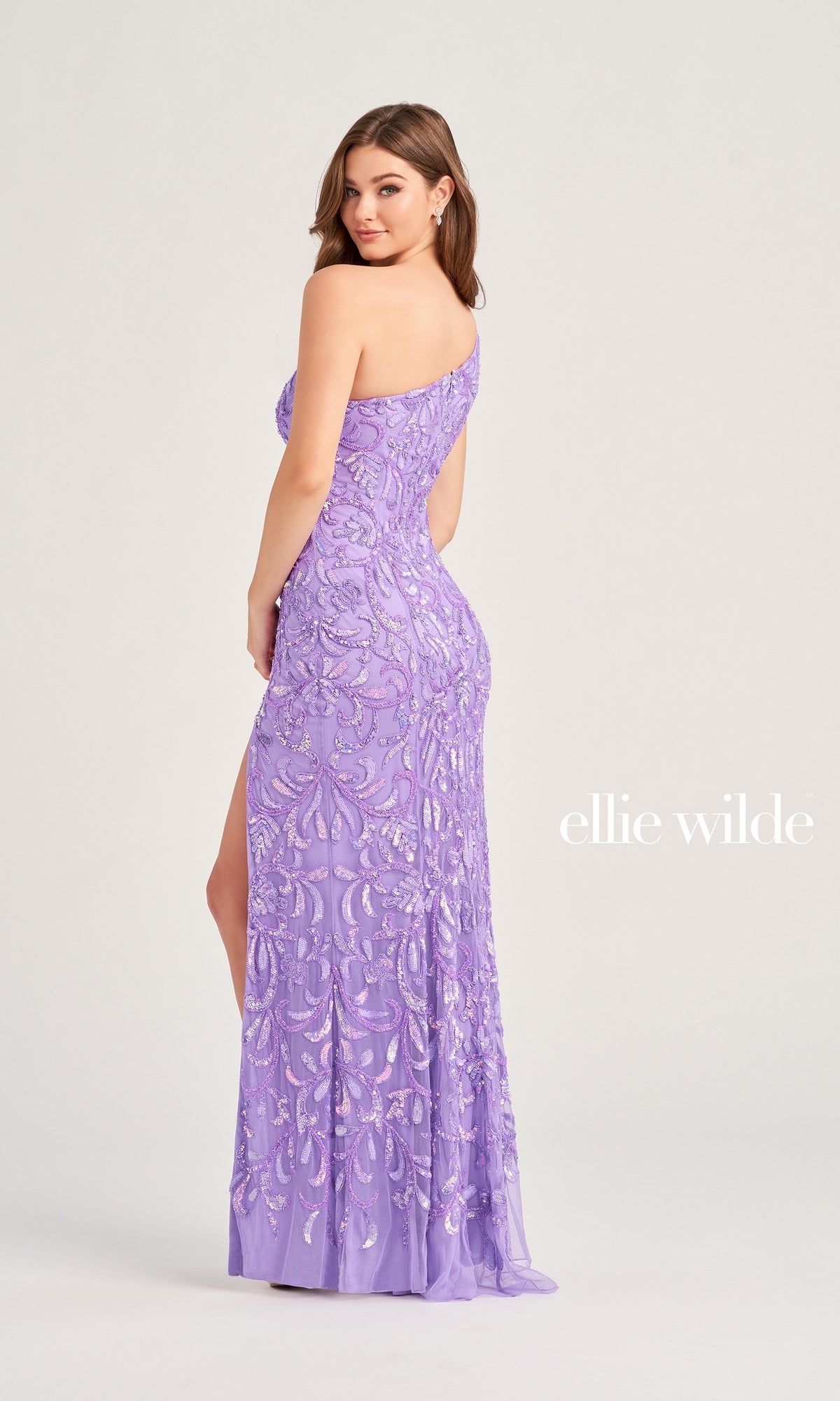 A woman models the Ellie Wilde One-Shoulder Beaded Prom Dress EW35021, featuring a light purple hue, sequin patterns, and a high slit. “ellie wilde” appears on the right against a plain background.